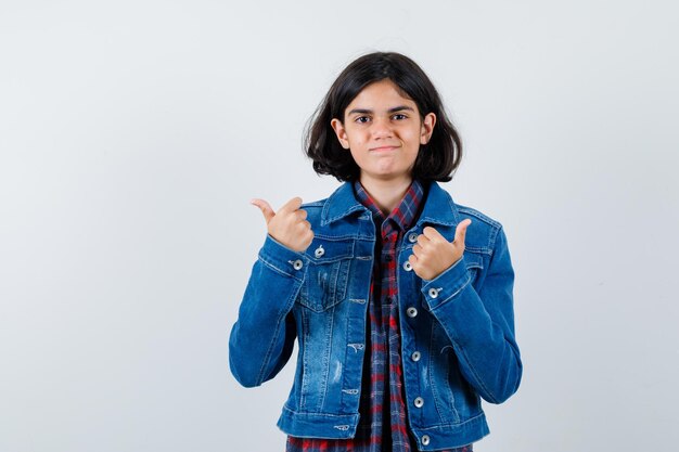 Giovane ragazza che mostra i pollici in su con entrambe le mani in camicia a quadri e giacca di jeans e sembra felice, vista frontale.