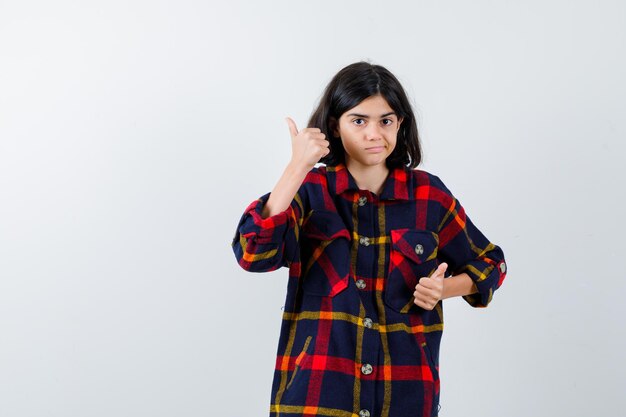 Giovane ragazza che mostra i pollici in su con entrambe le mani in camicia a quadri e sembra carina. vista frontale.