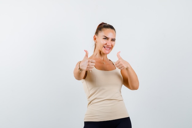 Giovane ragazza che mostra il doppio pollice in alto, ammiccante in top beige, pantaloni neri e guardando fiducioso. vista frontale.