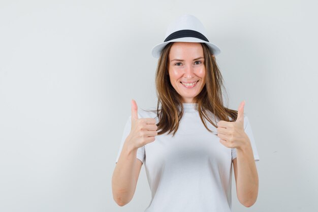 Giovane ragazza che mostra il doppio pollice in alto in t-shirt bianca, cappello e guardando allegro, vista frontale.