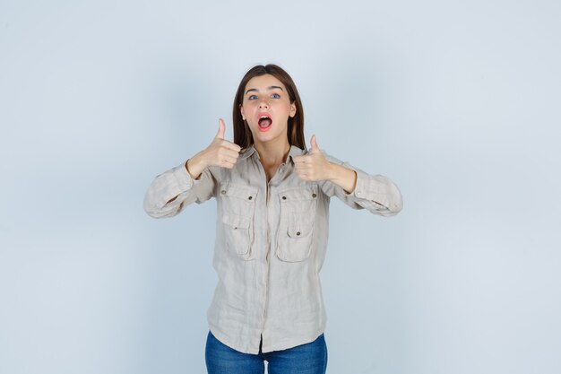 Giovane ragazza che mostra il doppio pollice in alto, tenendo la bocca spalancata in camicia beige, jeans e sembra carina, vista frontale.