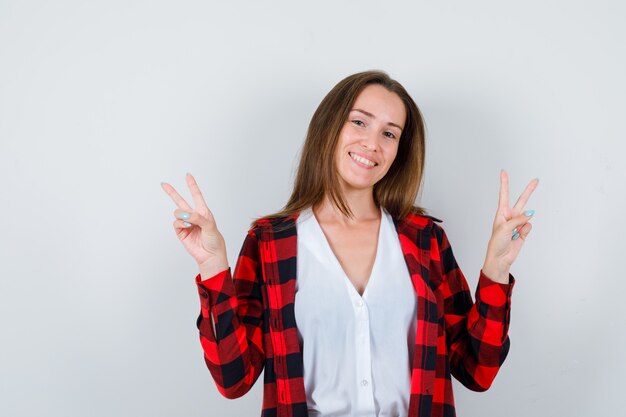 Giovane ragazza che mostra il doppio segno a V in camicia a scacchi, camicetta e sembra soddisfatta, vista frontale.