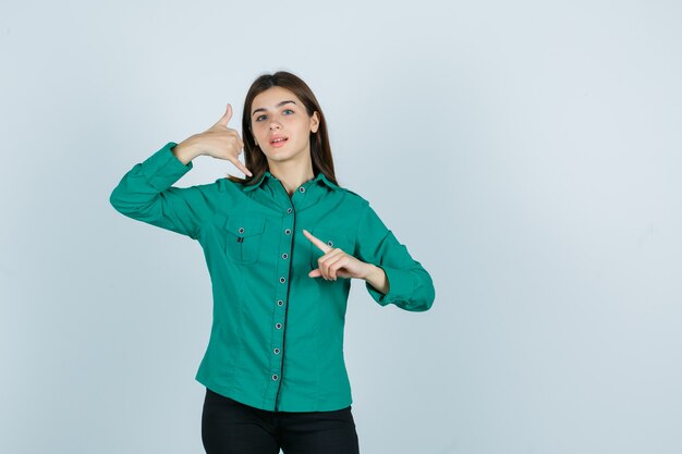 Giovane ragazza che mostra il gesto del telefono, indicando il lato sinistro in camicetta verde, pantaloni neri e guardando sanguigno. vista frontale.