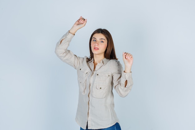 Giovane ragazza che mostra il gesto del vincitore in camicia beige, jeans e sembra fortunata. vista frontale.