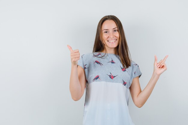 Giovane ragazza che mostra il pollice in alto mentre punta lontano in t-shirt e sembra allegro, vista frontale.