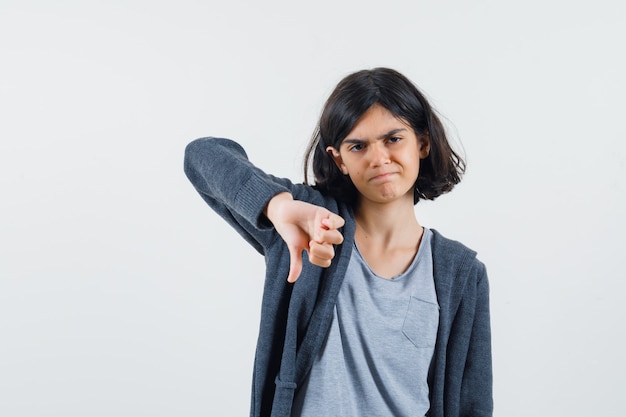 Giovane ragazza che mostra il pollice verso il basso in t-shirt grigio chiaro e felpa con cappuccio grigio scuro con zip e sembra scontenta