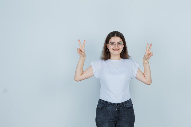 Giovane ragazza che mostra il segno di vittoria in t-shirt, jeans e che sembra felice, vista frontale.