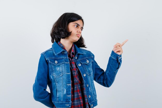 Giovane ragazza che punta a destra con il dito indice in camicia a quadri e giacca di jeans e sembra calma. vista frontale.