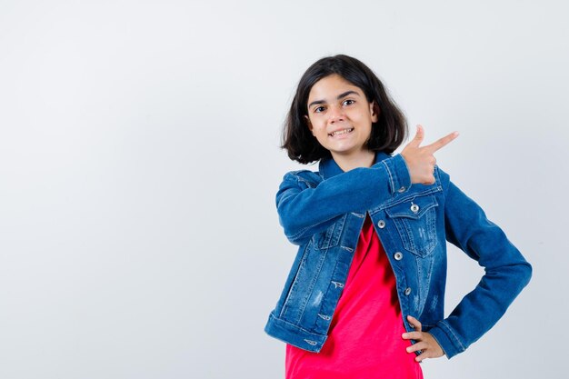 Giovane ragazza che punta a destra mentre tiene la mano sulla vita in maglietta rossa e giacca di jeans e sembra felice.