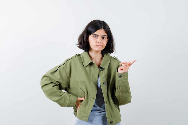 Giovane ragazza che punta a destra mentre tiene la mano sulla vita in maglione grigio, giacca kaki, pantaloni jeans e sembra seria, vista frontale.