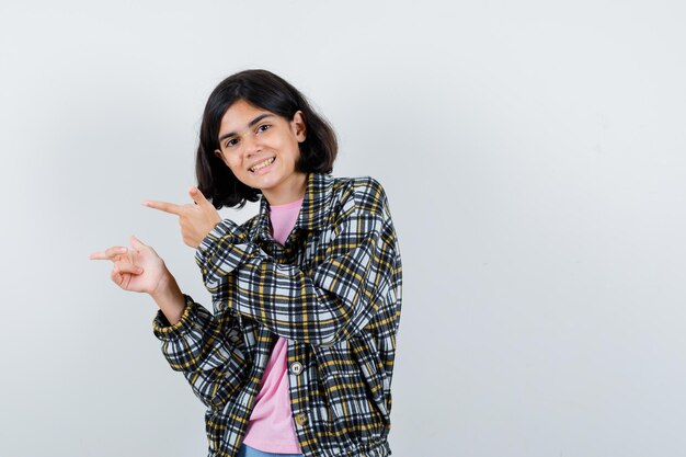 Giovane ragazza che punta a sinistra con l'indice in camicia a quadri e t-shirt rosa e sembra carina. vista frontale.