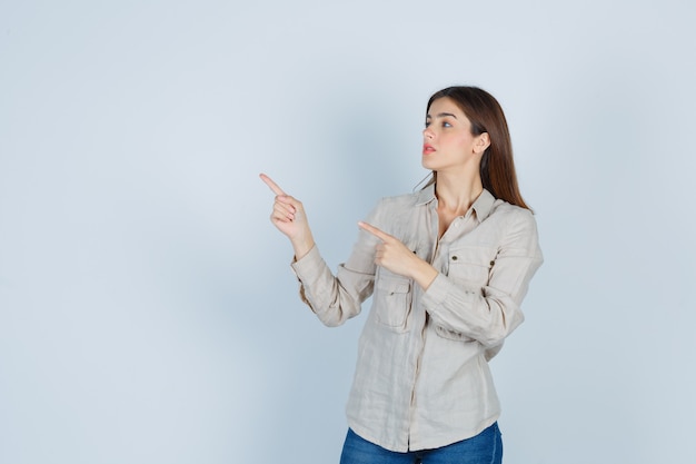 Giovane ragazza che punta a sinistra con le dita indice, guardando il lato sinistro in camicia beige, jeans e guardando a fuoco. vista frontale.