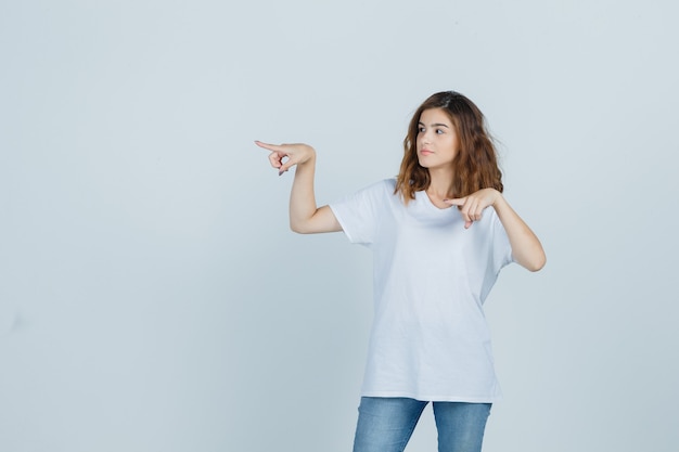 Giovane ragazza che punta a sinistra in t-shirt, jeans e guardando fiducioso, vista frontale.