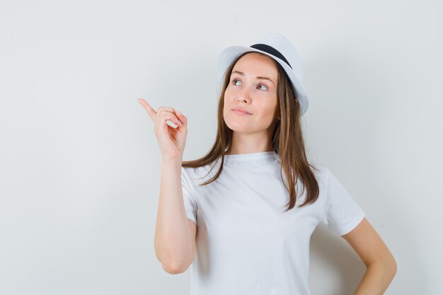 Giovane ragazza che punta all'angolo superiore sinistro in maglietta bianca, cappello e guardando fiducioso, vista frontale.