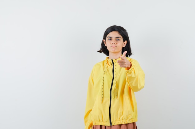 Giovane ragazza che punta alla telecamera con il dito indice in bomber giallo e gonna rosa e sembra seria.