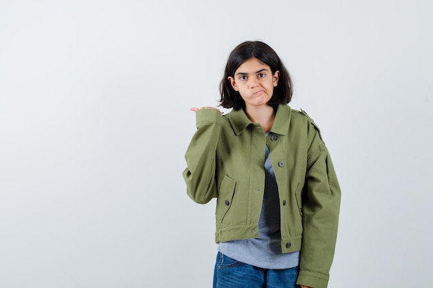 Giovane ragazza che punta lontano, incurvando le labbra in maglione grigio, giacca color kaki, pantaloni di jeans e sembra carina, vista frontale.