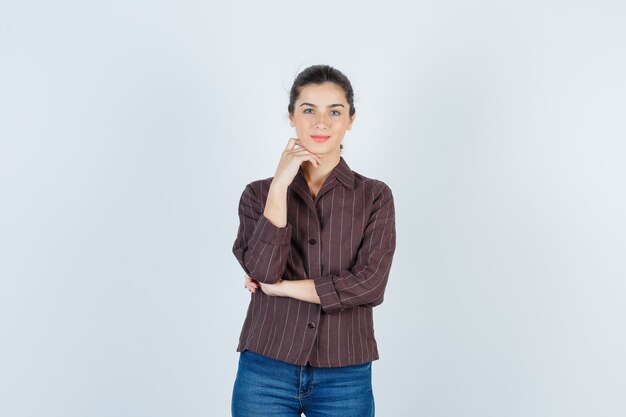 Giovane ragazza che puntella il mento a portata di mano in camicia a righe, jeans e sembra felice. vista frontale.