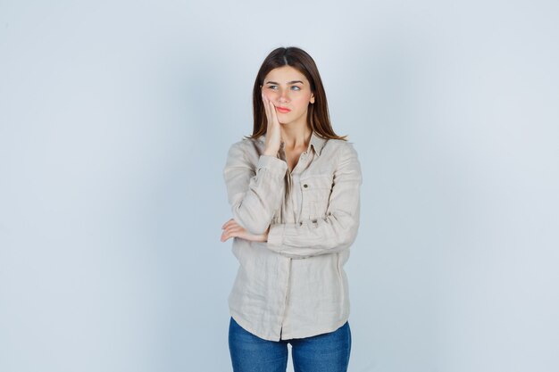 Giovane ragazza che si appoggia guancia sul palmo, distoglie lo sguardo in camicia beige, jeans e sembra carina, vista frontale.