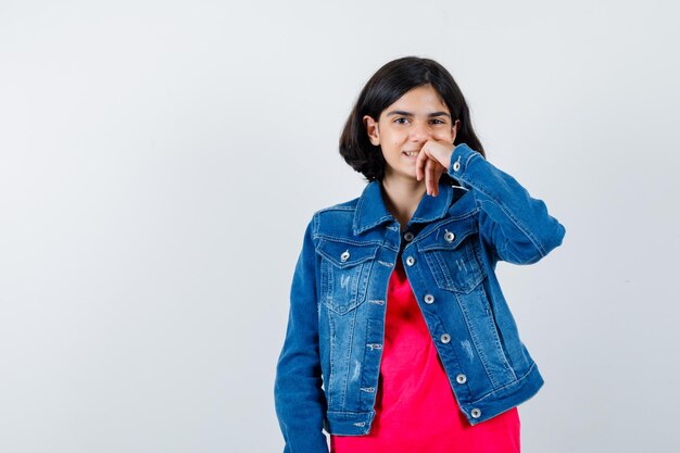 Giovane ragazza che si appoggia il mento sul palmo mentre pensa a qualcosa in maglietta rossa e giacca di jeans e sembra felice. vista frontale.