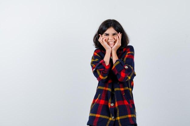 Giovane ragazza che si tiene per mano sulle guance in camicia a quadri e sembra felice, vista frontale.