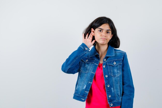 Giovane ragazza che si tiene per mano vicino all'orecchio per sentire qualcosa in maglietta rossa e giacca di jeans e sembra concentrata, vista frontale.