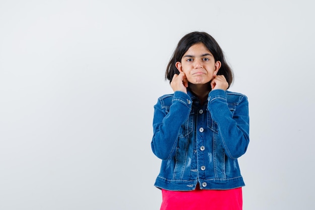 Giovane ragazza che si tiene per mano vicino all'orecchio per sentire qualcosa in maglietta rossa e giacca di jeans e sembra concentrata, vista frontale.