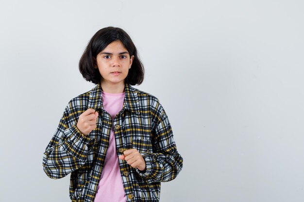 Giovane ragazza che stringe i pugni in camicia a quadri e t-shirt rosa e sembra arrabbiata, vista frontale.