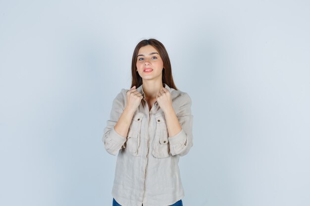 Giovane ragazza che stringe i pugni sul petto in camicia beige, jeans e sembra allegra. vista frontale.