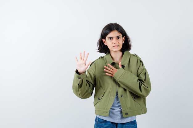 Giovane ragazza che tiene la mano sul petto e fa una promessa o un giuramento in maglione grigio, giacca kaki, pantaloni di jeans e sembra seria, vista frontale.