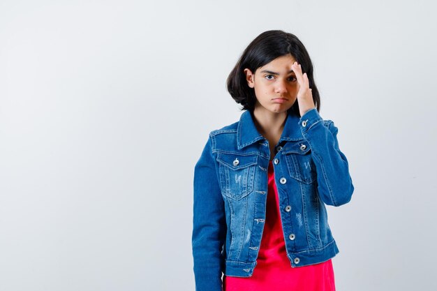 Giovane ragazza che tiene la mano sulla fronte in maglietta rossa e giacca di jeans e sembra seria, vista frontale.