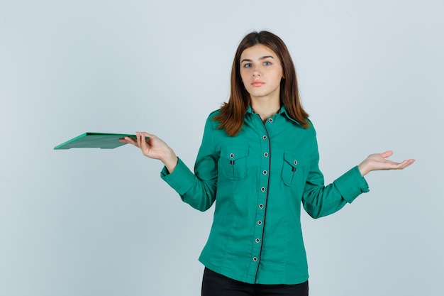 Giovane ragazza con appunti, mostrando gesto impotente in camicetta verde, pantaloni neri e guardando perplesso, vista frontale.