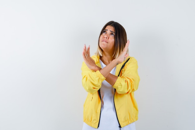 Giovane ragazza con due braccia incrociate, gesticolando nessun segno in t-shirt bianca, giacca gialla e guardando preoccupato, vista frontale.