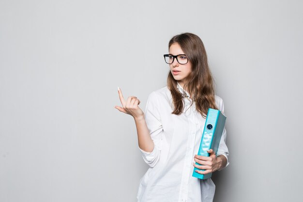 Giovane ragazza con gli occhiali vestita in t-shirt bianca da ufficio rigoroso si trova di fronte al muro bianco con la cartella blu per i documenti nelle sue mani