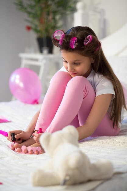 Giovane ragazza con i bigodini che si fa le unghie nella sua stanza. Piccola ragazza che indossa una maglietta bianca e leggins rosa seduta sul suo letto che si dipinge le unghie dei piedi.