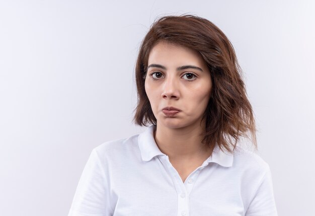 Giovane ragazza con i capelli corti che indossa la maglietta polo bianca che guarda l'obbiettivo accigliato e scontento