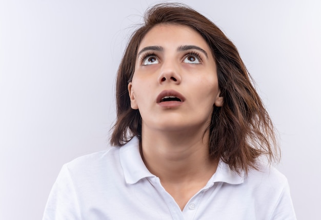 Giovane ragazza con i capelli corti che indossa la maglietta polo bianca che osserva in su sorpreso