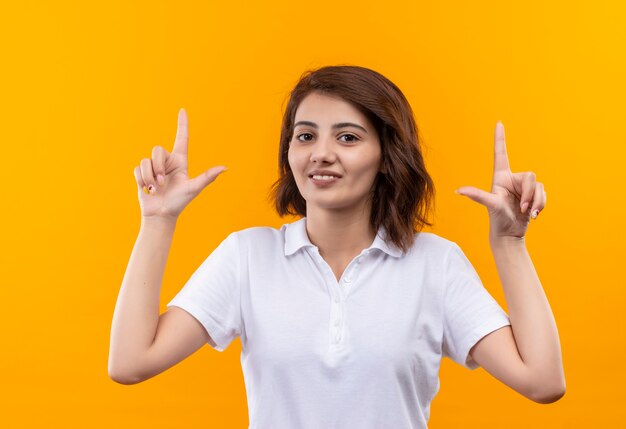 Giovane ragazza con i capelli corti che indossa la maglietta polo bianca guardando la telecamera sorridendo allegramente rivolta verso l'alto con il dito indice