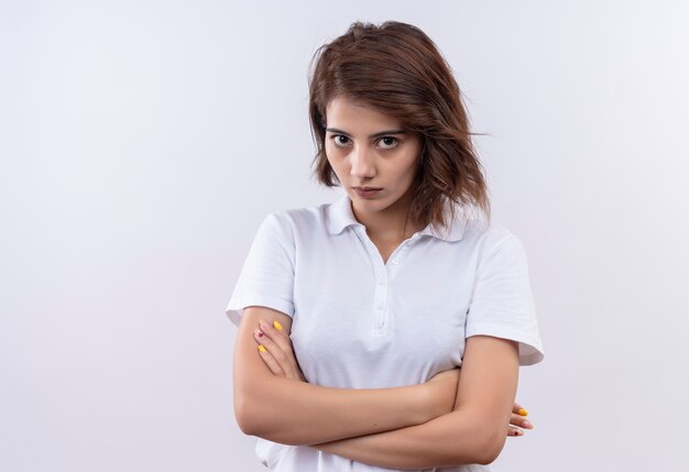 Giovane ragazza con i capelli corti che indossa la maglietta polo bianca in piedi con le mani incrociate sul petto guardando la telecamera displesed su sfondo bianco