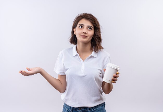 Giovane ragazza con i capelli corti che indossa la maglietta polo bianca tenendo la tazza di caffè cercando confuso diffondendo il braccio a lato