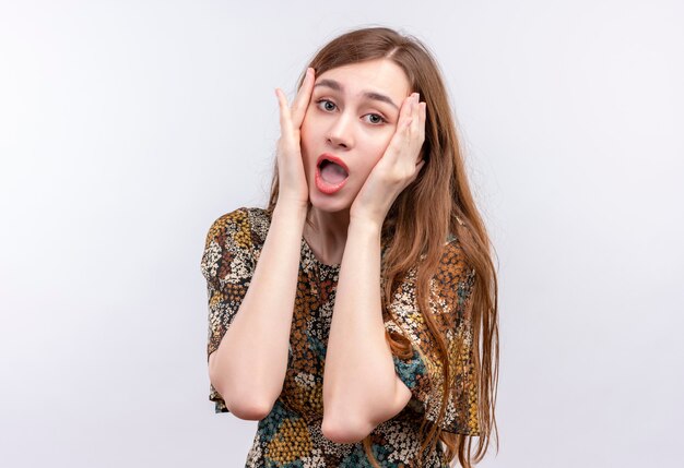 Giovane ragazza con i capelli lunghi che indossa abiti colorati guardando sorpreso e stupito toccando con le braccia il viso