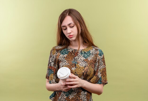 Giovane ragazza con i capelli lunghi che indossa abiti colorati tenendo la tazza di caffè guardandolo con espressione triste sul viso