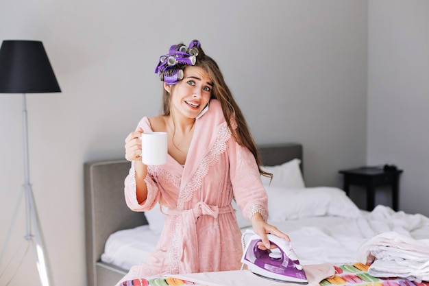 Giovane ragazza con i capelli lunghi in accappatoio rosa e bigodino sulla testa tenendo il ferro e parlando al telefono a casa. Sembra stupita.