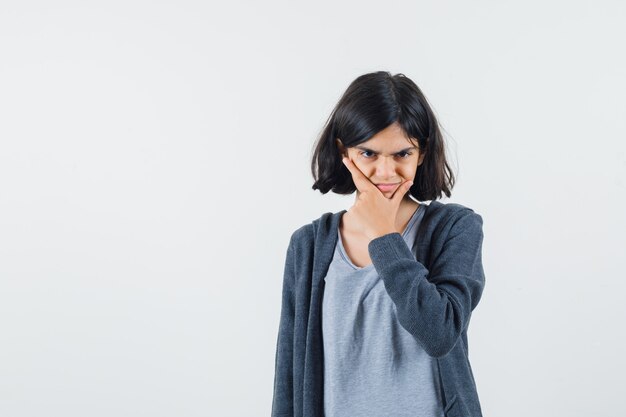 Giovane ragazza con il mento appoggiato sul palmo in t-shirt grigio chiaro e felpa con cappuccio con zip grigio scuro e dall'aria seria