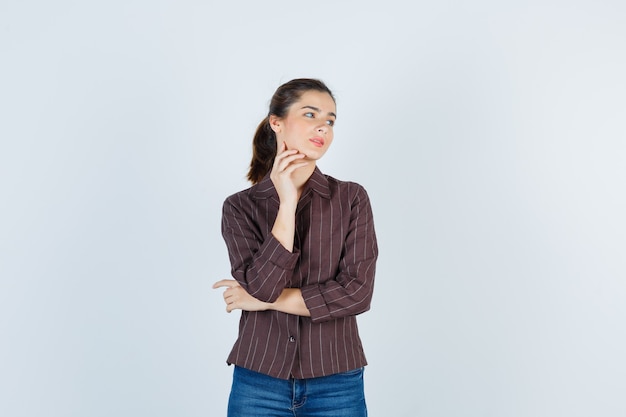 Giovane ragazza con la mano sulla guancia, guardando il lato destro in camicia a righe, jeans e guardando focalizzato, vista frontale.