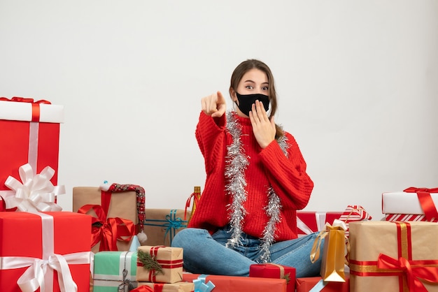 giovane ragazza con maglione rosso mettendo la mano sulla bocca che punta con il dito davanti seduto intorno presenta con maschera nera su bianco