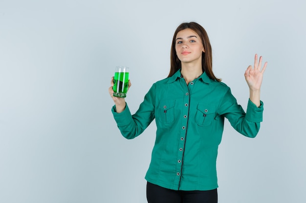 Giovane ragazza con un bicchiere di liquido verde, mostrando segno giusto in camicetta verde, pantaloni neri e guardando allegro. vista frontale.