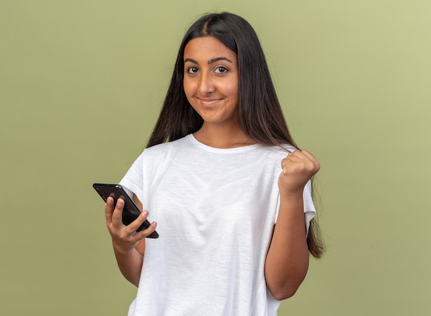 Giovane ragazza con una maglietta bianca che tiene in mano uno smartphone che guarda la telecamera pugno serrato felice ed eccitato