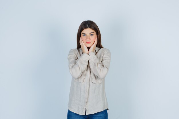 Giovane ragazza cuscino viso sulle mani in camicia beige, jeans e guardando piuttosto, vista frontale.