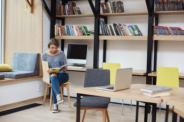 Giovane ragazza dai capelli chiari intelligente con acconciatura bob in abiti casual, seduta sulla sedia in biblioteca moderna, leggendo il libro preferito, rilassarsi dopo una lunga giornata di studio