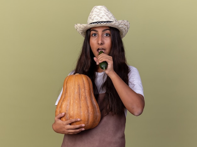 Giovane ragazza del giardiniere in grembiule e cappello estivo che tiene il cetriolo e la zucca con la faccia seria che sta sopra la parete verde